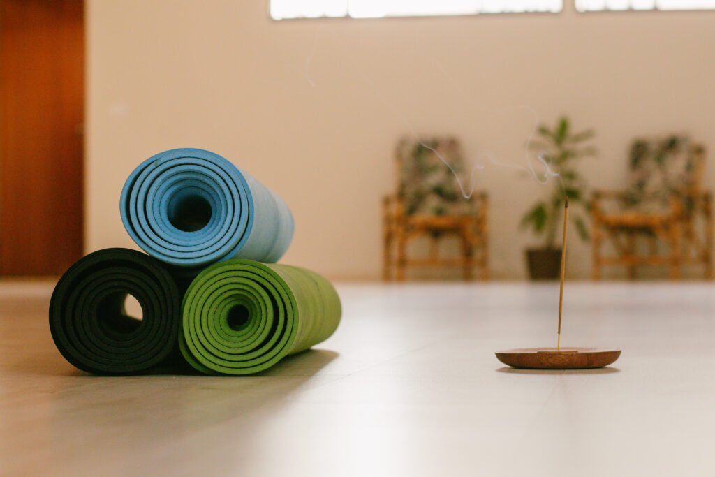 Três tapetes de yoga enrolados (verde, azul, preto) e um incenso aceso em um estúdio de yoga em Cianorte-PR, criando uma atmosfera calma.