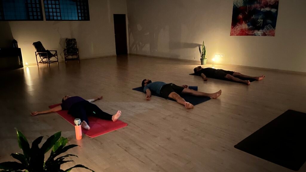 Três alunos deitados em Savasana (postura do cadáver) durante o relaxamento final de uma aula de yoga noturna em um estúdio em Cianorte-PR.