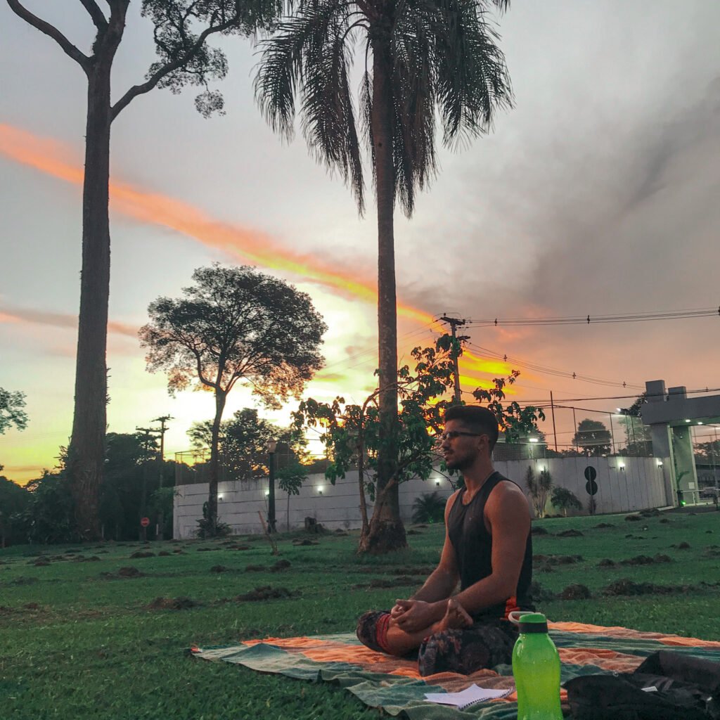 Vitor Lanaro praticando yoga sentado na grama em um parque de Cianorte, com um pôr do sol colorido ao fundo entre as árvores.