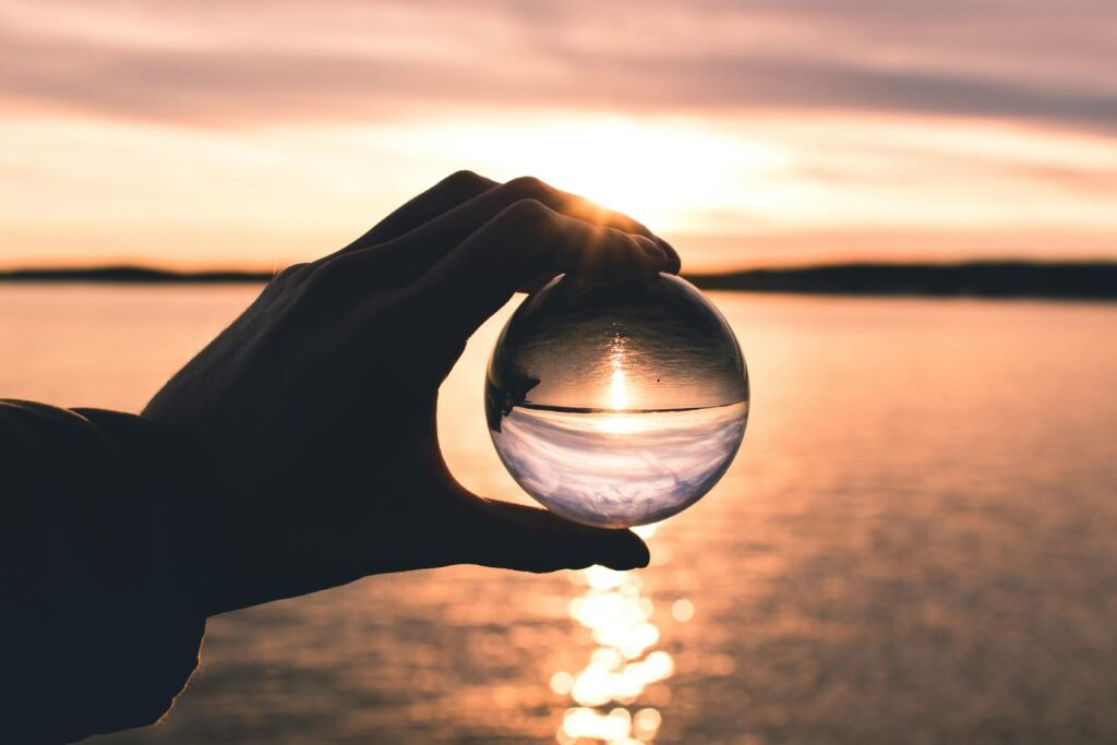 A hand holds a crystal ball reflecting a serene sunset over calm ocean waters.